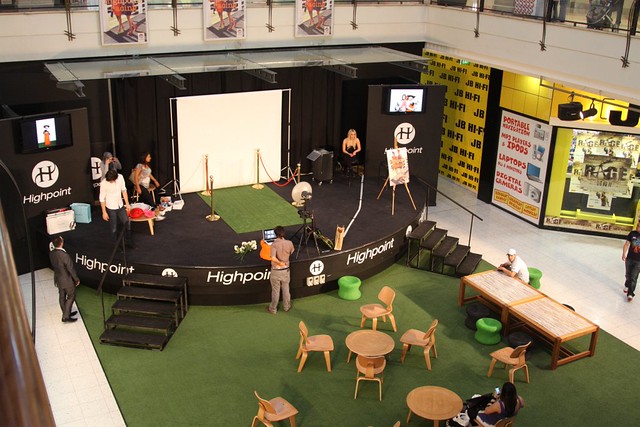 A portable black stage set up on green turf carpet with Highpoint branding, JB Hi-Fi signage visible in the background, chairs and tables arranged nearby