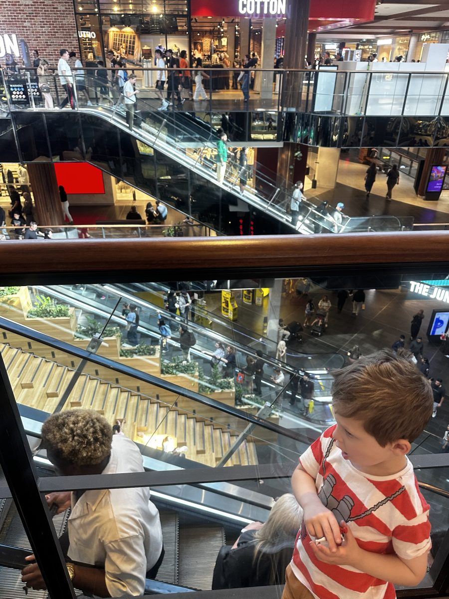 A young boy leaning on a glass balustrade at Highpoint, looking down at the escalators and timber steps below