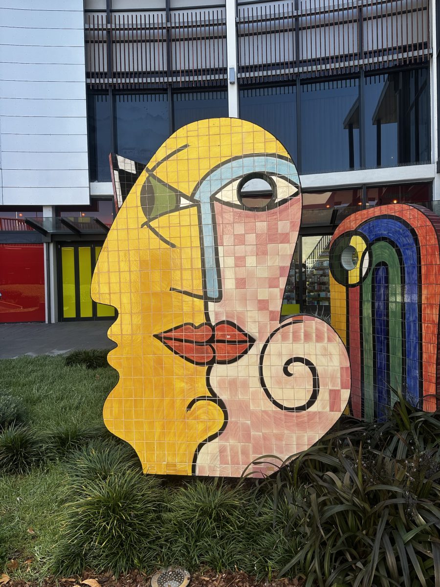 A tiled mosaic face sculpture in yellow and pink, with a rainbow-striped companion piece behind it