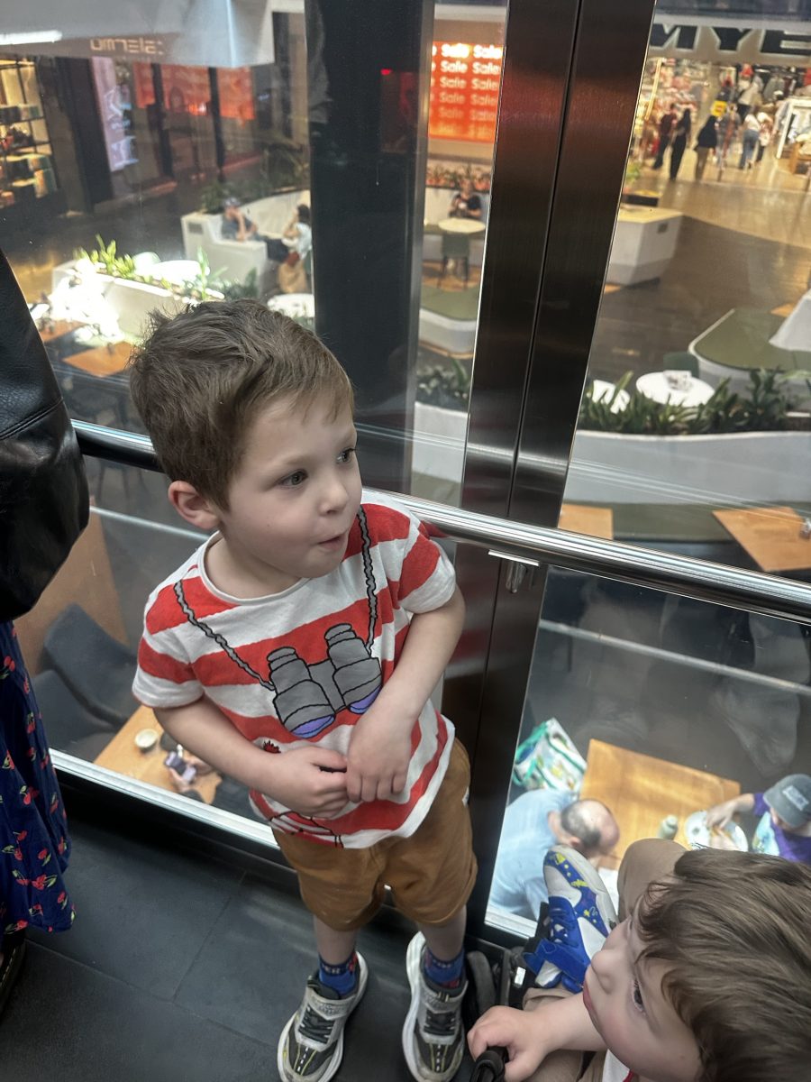 A young boy standing inside Highpoint's glass elevator, looking out through the glass towards the shopping centre and Myer signage