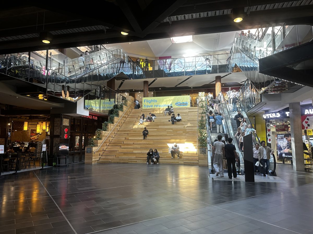 A wide timber amphitheatre-style staircase at Highpoint with people sitting on the steps, escalators to the right and Dumplings Plus to the left