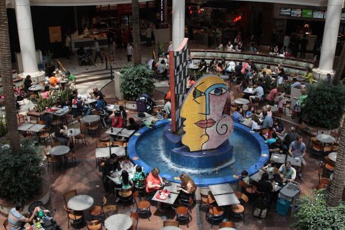 The Picasso-style yellow and pink face sculpture standing in Highpoint's circular blue fountain, surrounded by food court tables and diners