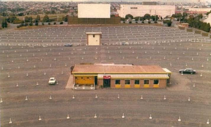 Aerial view of the Sunset Drive-In concession building and ticket booth, with the empty screen and rows of speaker posts stretching across the field