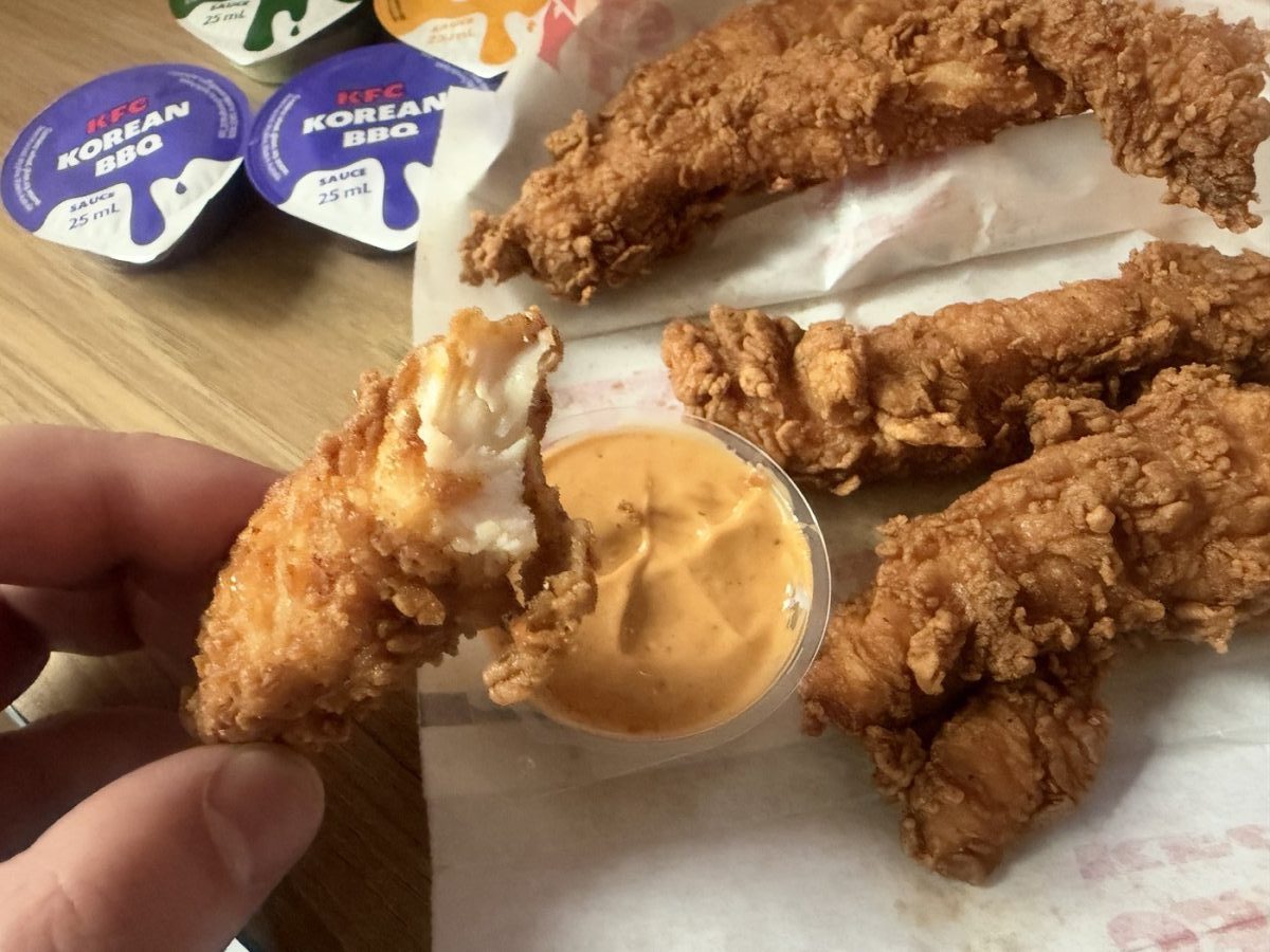 A bitten new KFC tender showing white chicken inside, held next to a tub of orange dipping sauce