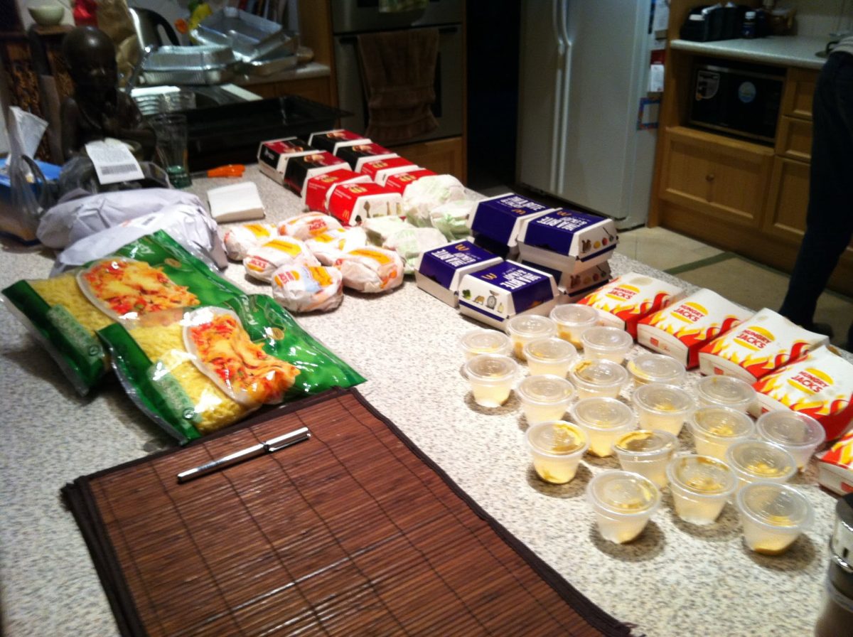 A kitchen bench covered in McDonald's and Hungry Jack's burgers, Hungry Jack's onion rings, bags of grated cheese, bacon in butcher's paper and around 20 clear plastic cups with small amounts of Big Mac sauce