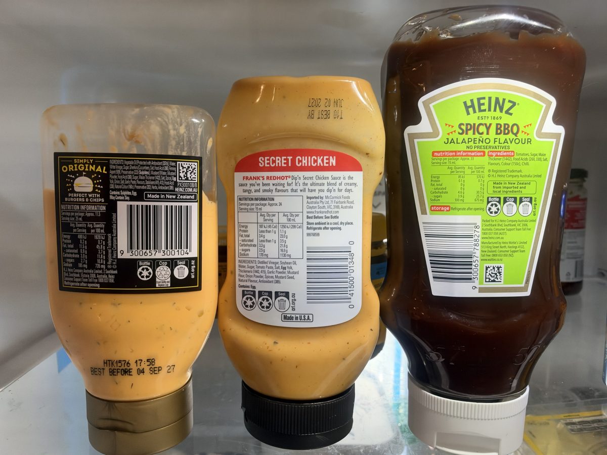 Three upside-down sauce bottles on a fridge shelf, Heinz Simply Original burger sauce, Frank's RedHot Secret Chicken and Heinz Spicy BBQ Jalapeño, each with a label saying to refrigerate after opening.